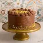 Dubai Chocolate Cake: An Irresistible Homemade Masterpiece 3 A luxurious Dubai chocolate cake decorated with piped chocolate swirls and crushed pistachios, displayed on a golden cake stand with a Moroccan-style tile background.