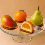 Hyper-realistic fruit shaped desserts including mango, orange, and pear pastries displayed on a glass stand.